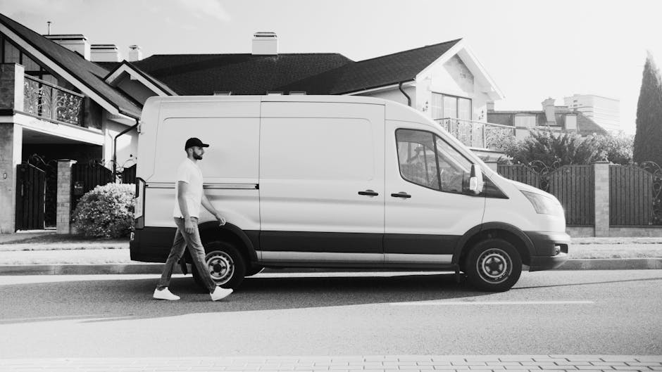 A man wearing a white T-shirt, dark pants, and a baseball cap is walking alongside a white delivery van parked on a residential street. The van is positioned parallel to the curb, with its sliding sid